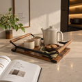 A wooden serving tray with black metal frame and light wood handles, styled on a marble countertop with a ceramic teapot, two cups, a small bowl of cookies, and a folded green cloth. The scene is set in a warm, modern kitchen environment.