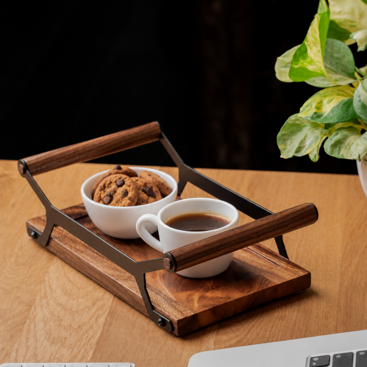 A wooden coffee tray with dark metal handles sits on a wooden desk, holding a white cup of coffee and a white bowl of cookies. A potted plant and part of a keyboard are visible in the scene.