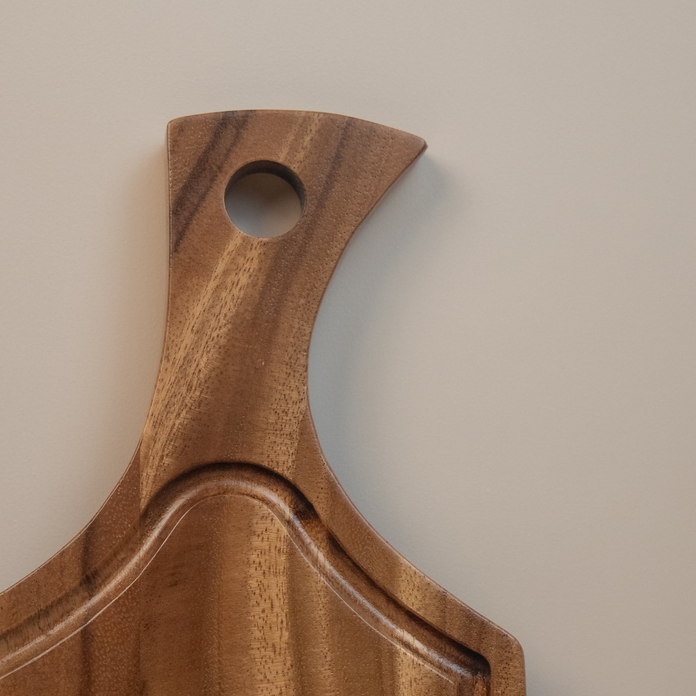 A close-up of a wooden serving board or tray with a curved handle and a circular cutout, showing a warm medium-brown wood grain against a plain light background.