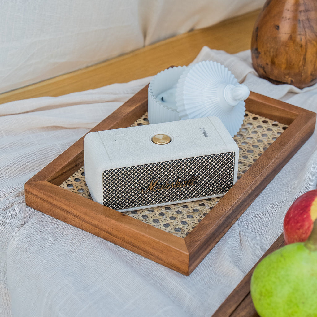 A white Marshall portable speaker sits on a wooden tray with a woven mat, styled on draped white fabric with a wooden vase and fruit in the background.