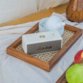 A white Marshall portable speaker sits on a wooden tray with a woven mat, styled on draped white fabric with a wooden vase and fruit in the background.