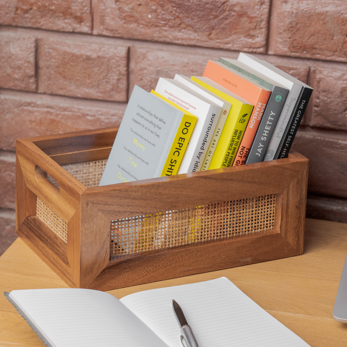 A wooden storage crate with woven cane panels sits on a wooden table, holding several books. An open notebook and pen are in the foreground, with a brick wall background.