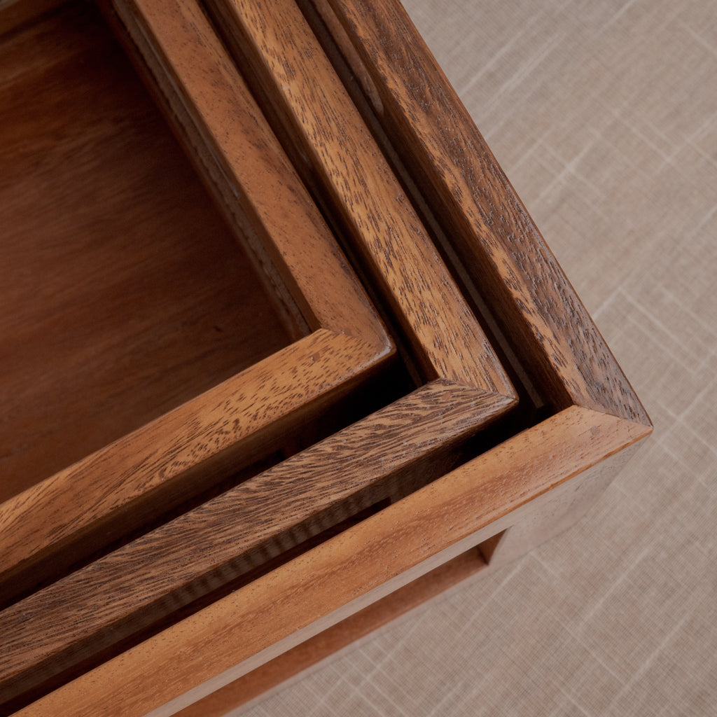 Close-up of a wooden nesting cabinet or tray set with warm medium-brown wood grain and a woven cane insert, shown on a beige textured floor.