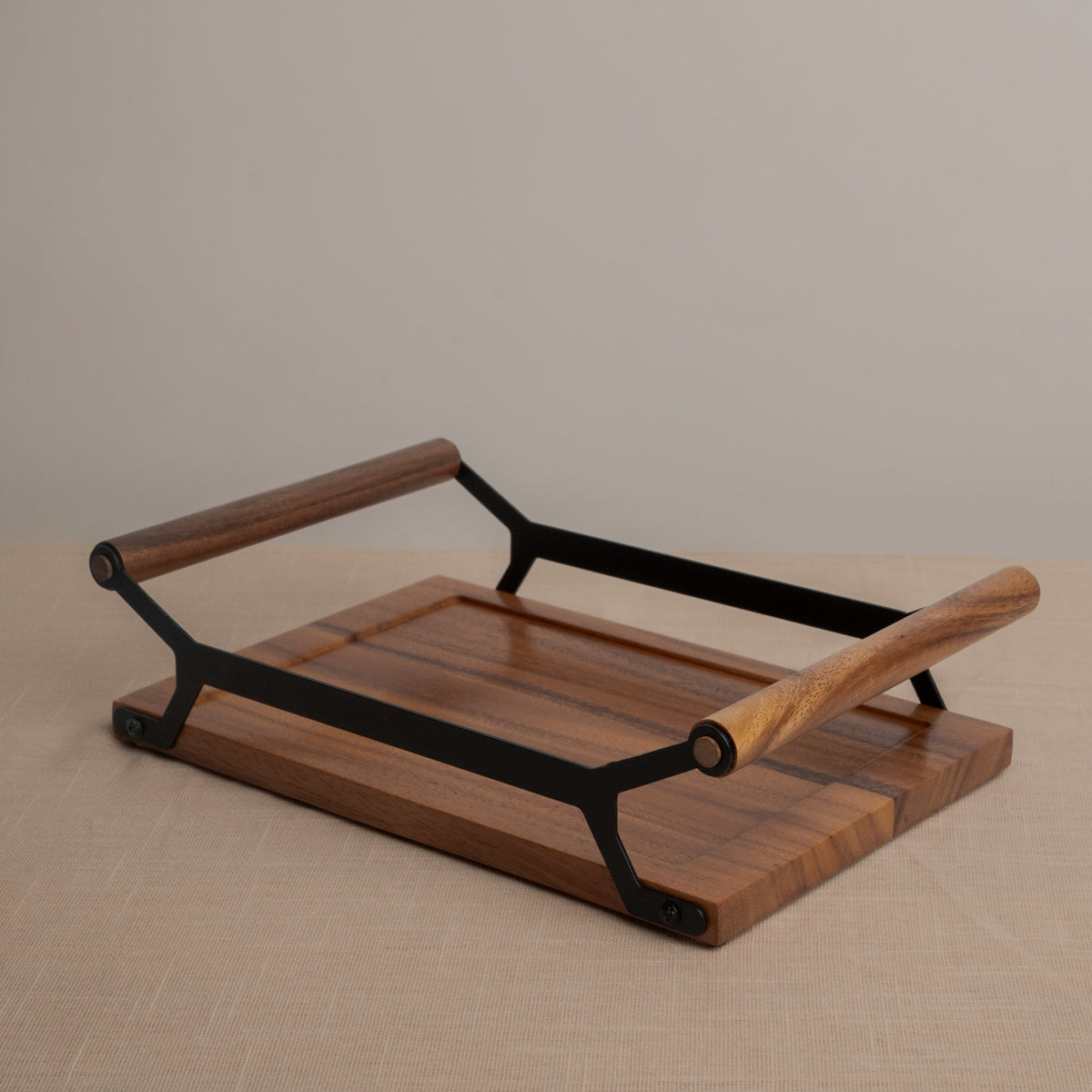 A rectangular wooden serving tray with a dark brown wood grain finish and black metal frame supports two wooden handles, photographed on a neutral beige textured surface against a light gray background.