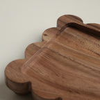 A close-up of a scalloped wooden serving tray with a smooth, slightly glossy finish and visible wood grain, set against a neutral light gray background.