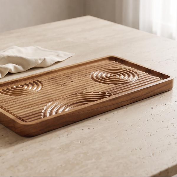 A rectangular wooden tray with carved circular wave patterns and a slotted top, styled on a light stone tabletop beside a small bowl and draped fabric.