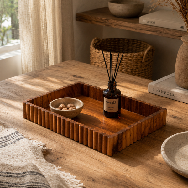 A rectangular wooden tray with a fluted edge sits on a rustic wooden table in a warm, sunlit room. The tray holds a small bowl of pebbles and a dark glass reed diffuser bottle.
